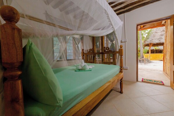 A bed with a green sheet and a wooden headboard, a white canopy, and a view of an outdoor area with a thatched structure.