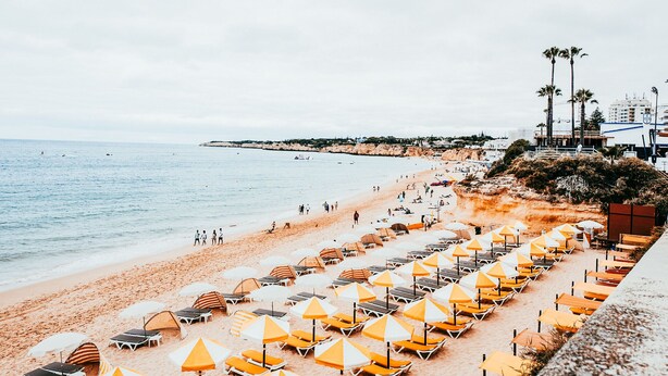 A beach with rows of sun loungers and umbrellas, a sandy shore, and a calm sea.