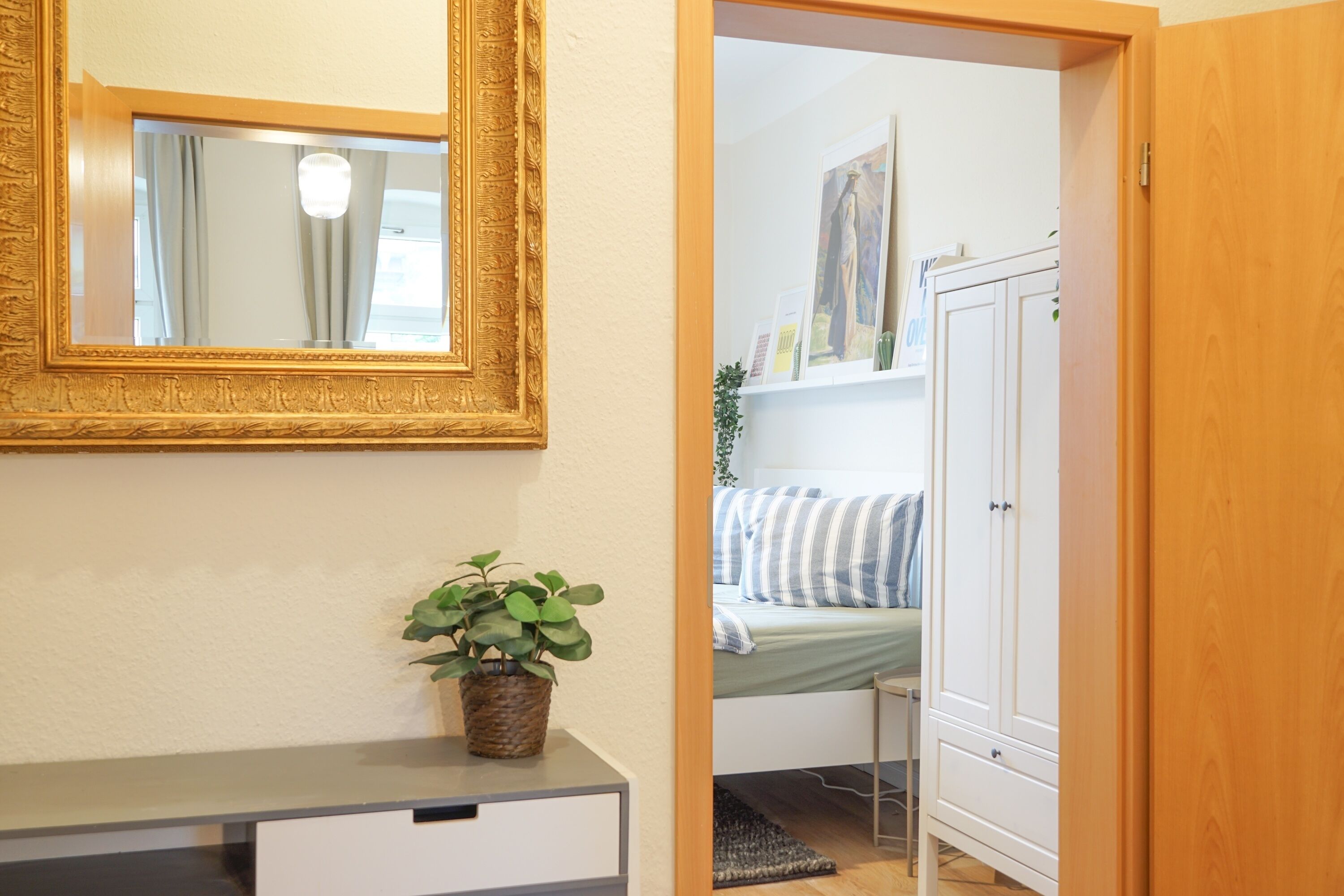 A room with a bed, a bedside table with a plant, a wardrobe, and a mirror on the wall.