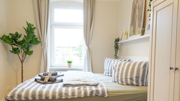 A neatly made bed with striped bedding, a bedside plant, and a window with curtains. - Cosy & quiet apartment w/great transport links (Erfurt)