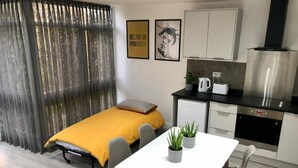 A compact room with a bed, a small kitchenette, a dining table, and a window with sheer curtains. - Cosy Flat for 1-3 not far From City Centre (Leicester)