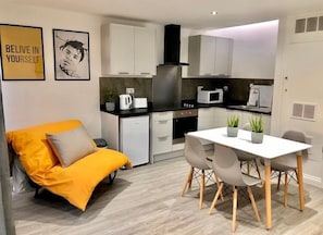 A compact kitchen with white cabinets, a microwave, and a small refrigerator. There is a dining table with chairs and a potted plant.