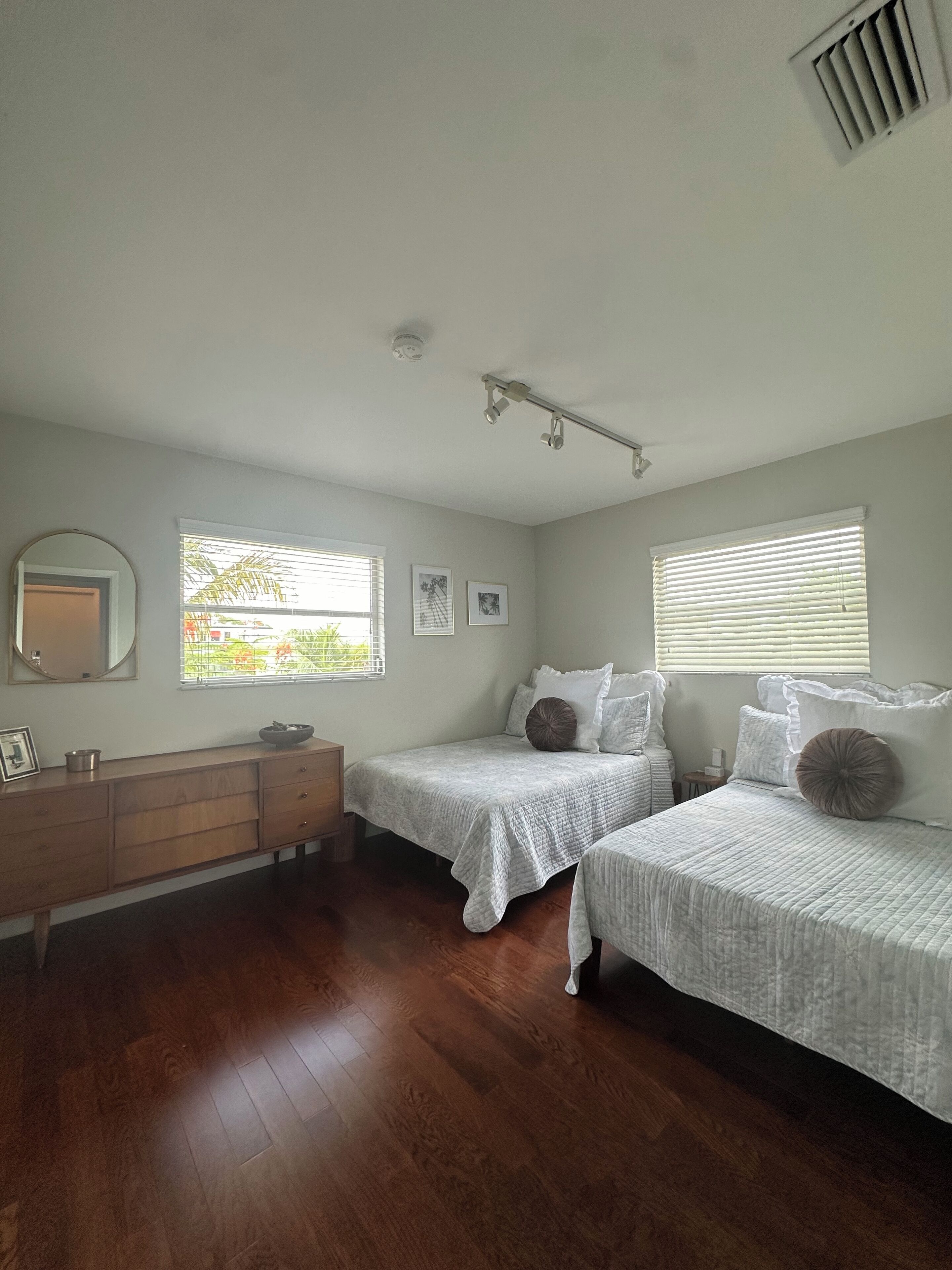 A bedroom with two beds, a dresser, a mirror, and two windows with blinds.