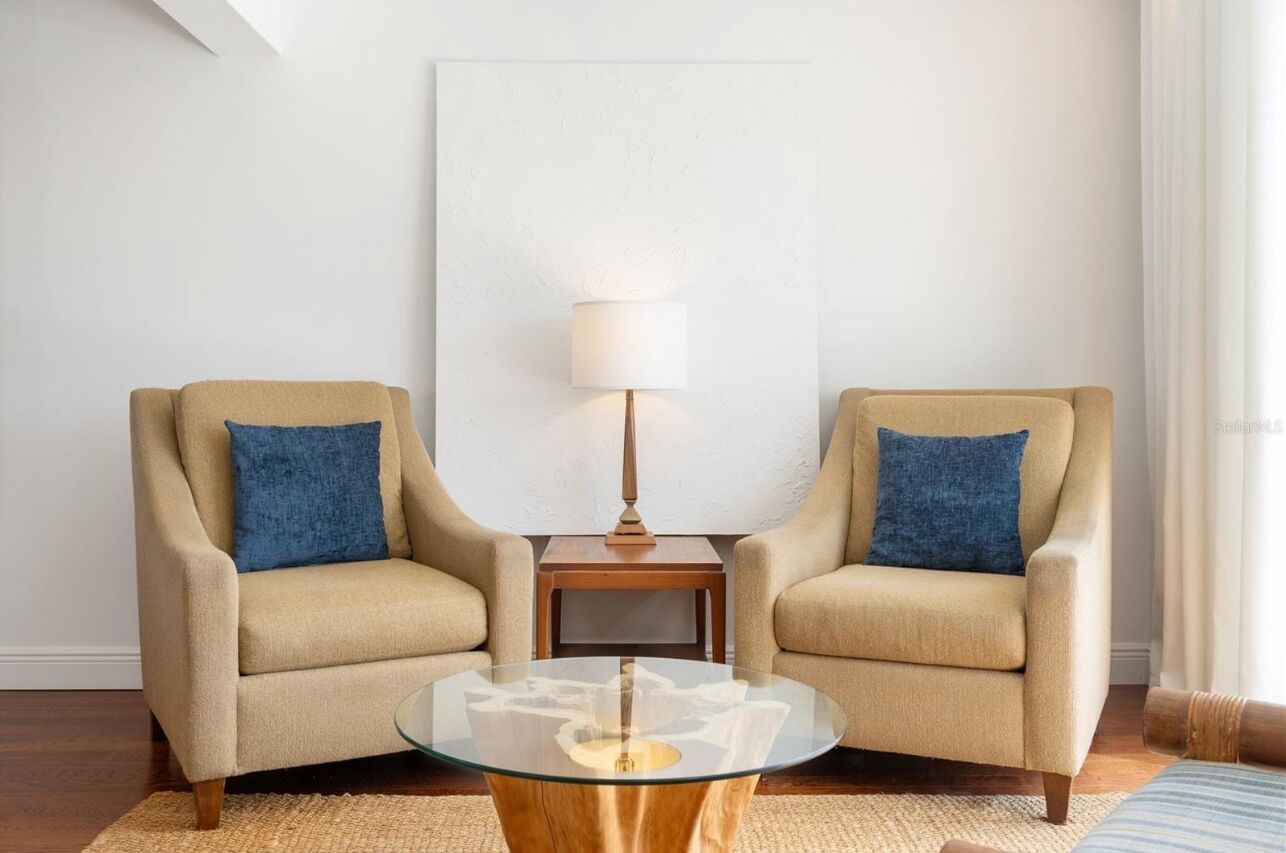 Una habitación con dos sillones color beige, una mesa de centro de cristal, una mesita de noche de madera con una lámpara, y un cuadro abstracto grande en la pared.