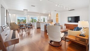 Een moderne woonkamer met een piano, een bank, een eethoek met stoelen, en een keuken op de achtergrond.