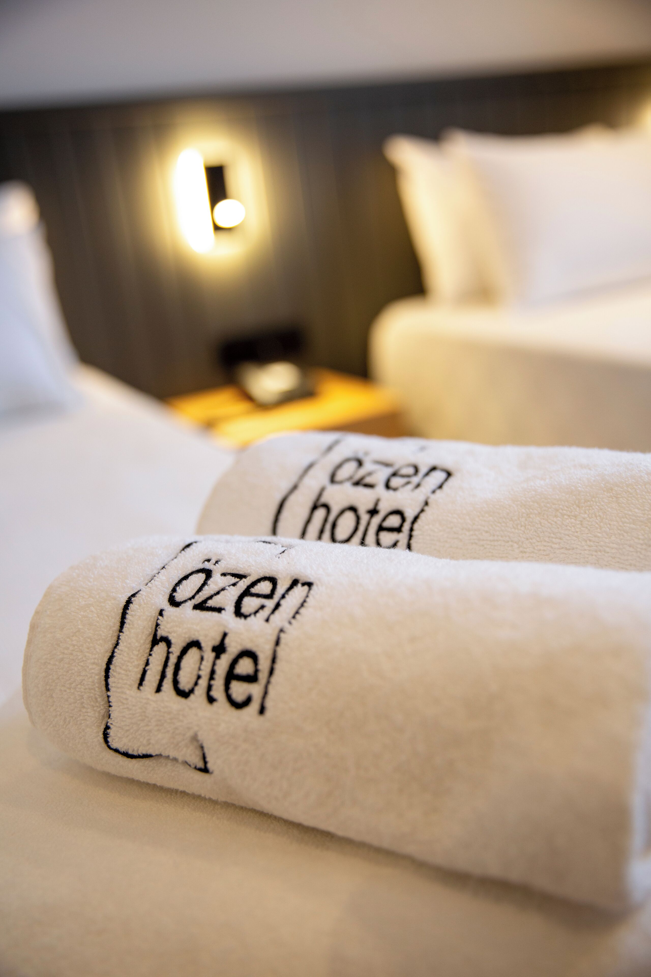 Two neatly folded towels with the text "ozen hotel" on them, placed on a bed with a headboard and a bedside table with a lamp.