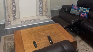 Ein Wohnzimmer mit Kamin, einem darüber montierten Fernseher, einem Ledersofa mit farbigem Kissen und zwei Fernbedienungen auf einem Holztisch.