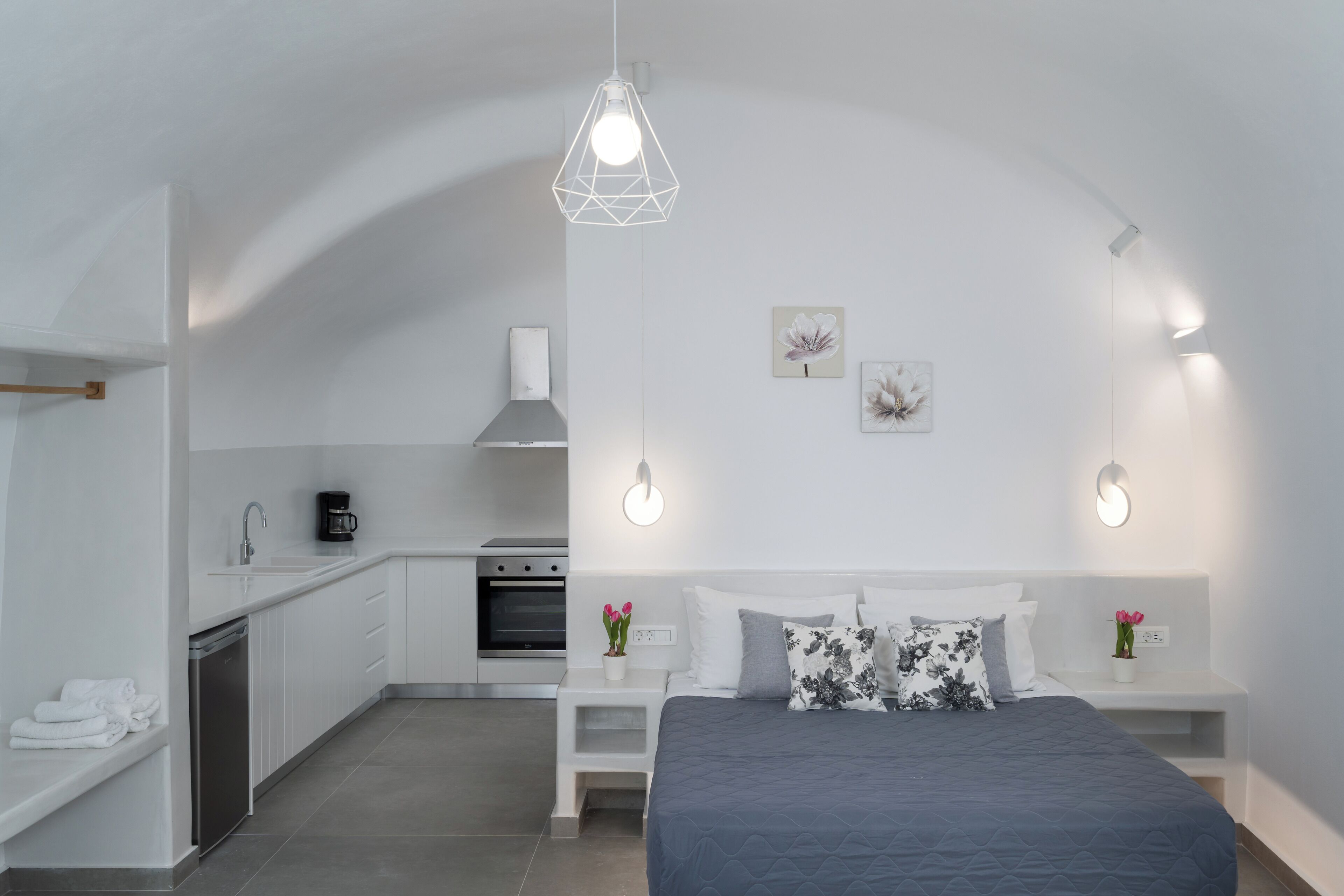 A modern bedroom with a large bed, bedside tables, and a kitchen area in the background.