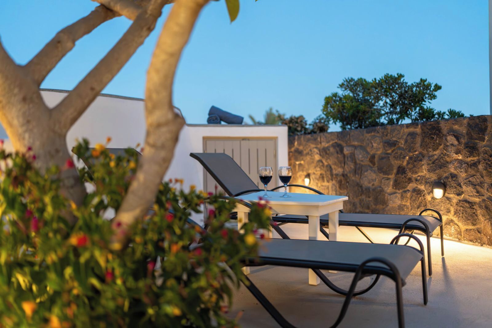 An outdoor seating area with two lounge chairs and a table, two wine glasses, and a stone wall in the background.