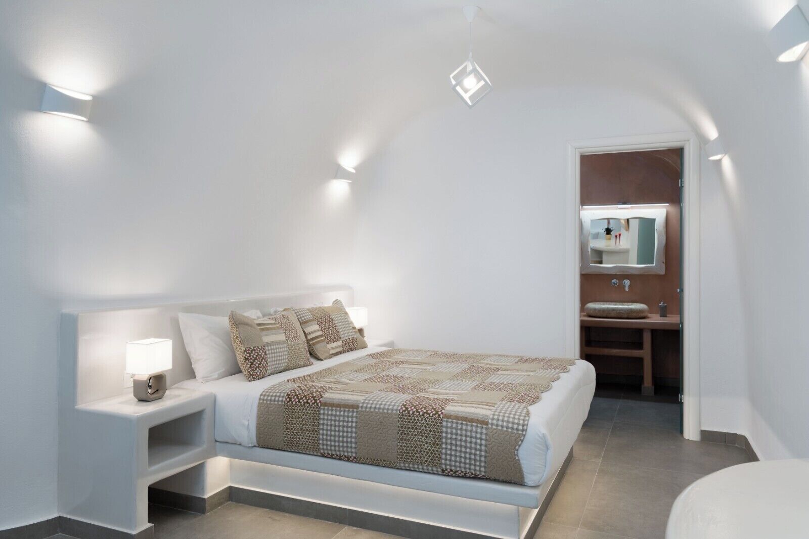 Un dormitorio con cama, mesita de noche y mesilla con lámpara. Se ve un baño con lavamanos y espejo.