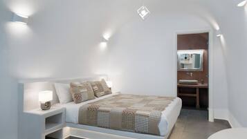 A bedroom with a bed, bedside table, and a nightstand with a lamp. There is a bathroom visible with a sink and mirror.