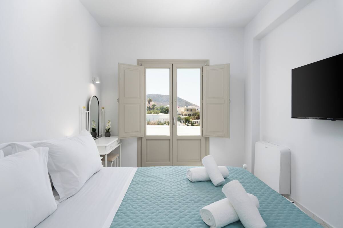 A hotel room with a bed, a TV mounted on the wall, and a view of a mountain through open doors.
