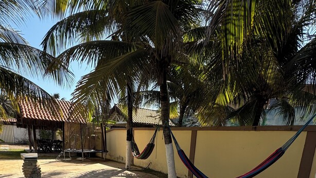 A swimming pool with a stone deck, surrounded by palm trees and a tiled-roof structure. - Home Beach Mar (Aquiraz)