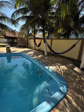A swimming pool with a stone deck, surrounded by palm trees and a tiled-roof structure. - Home Beach Mar (Aquiraz)