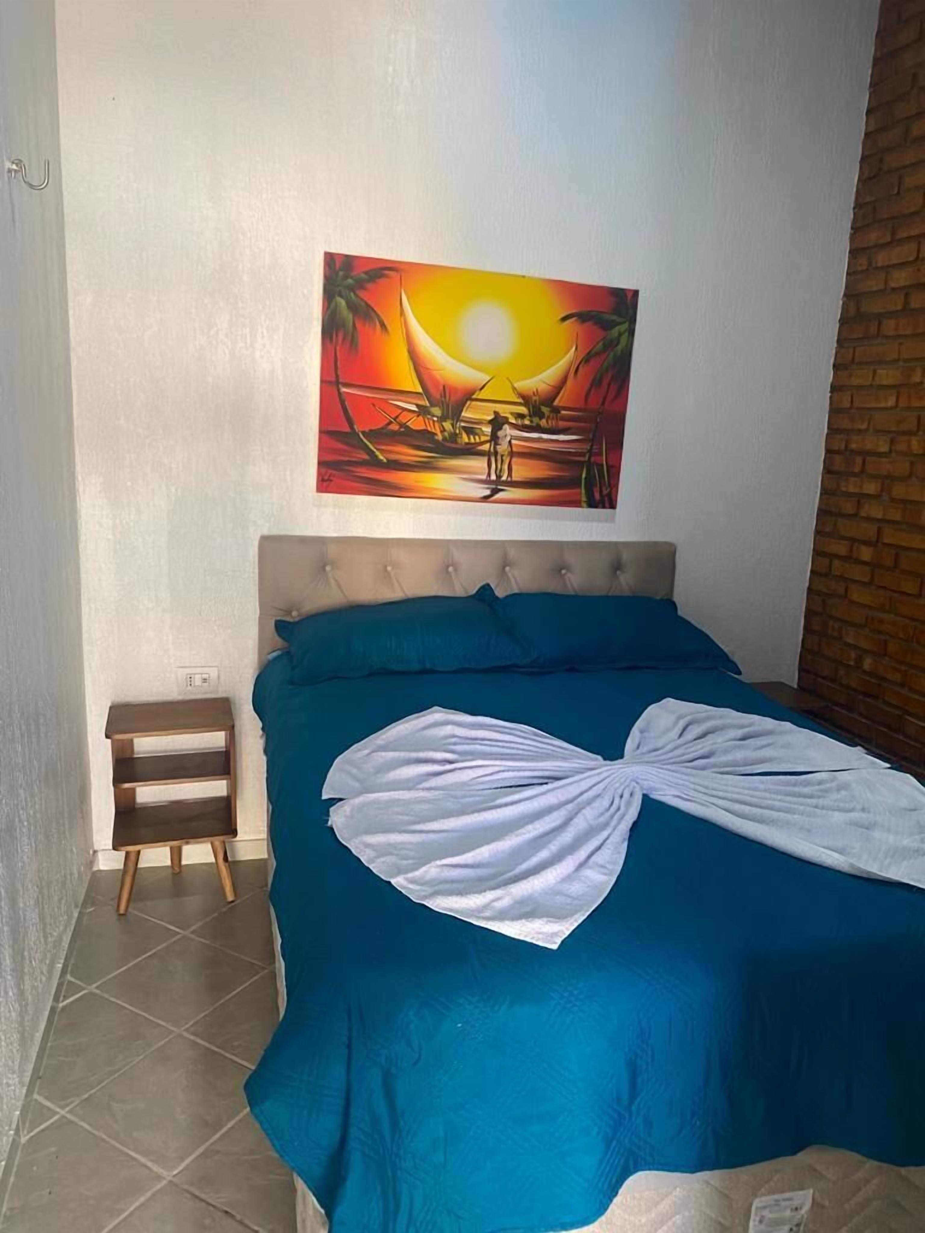 A bedroom with a bed, a nightstand, a painting of a sunset, and a brick wall.