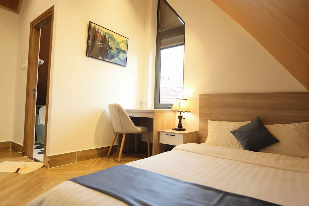 A bedroom with a bed, a desk, a chair, a nightstand with a lamp, a painting, and a window.