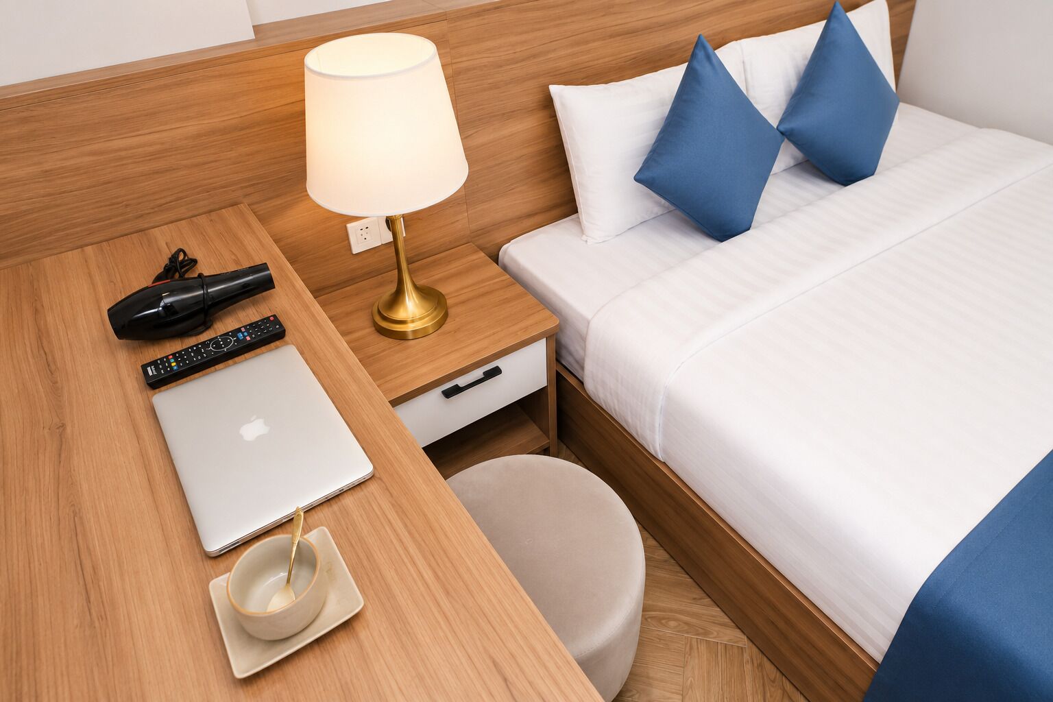 A hotel room with a wooden headboard, a bed with white bedding and blue pillows, a bedside table with a lamp, a laptop, a remote control, and a cup.
