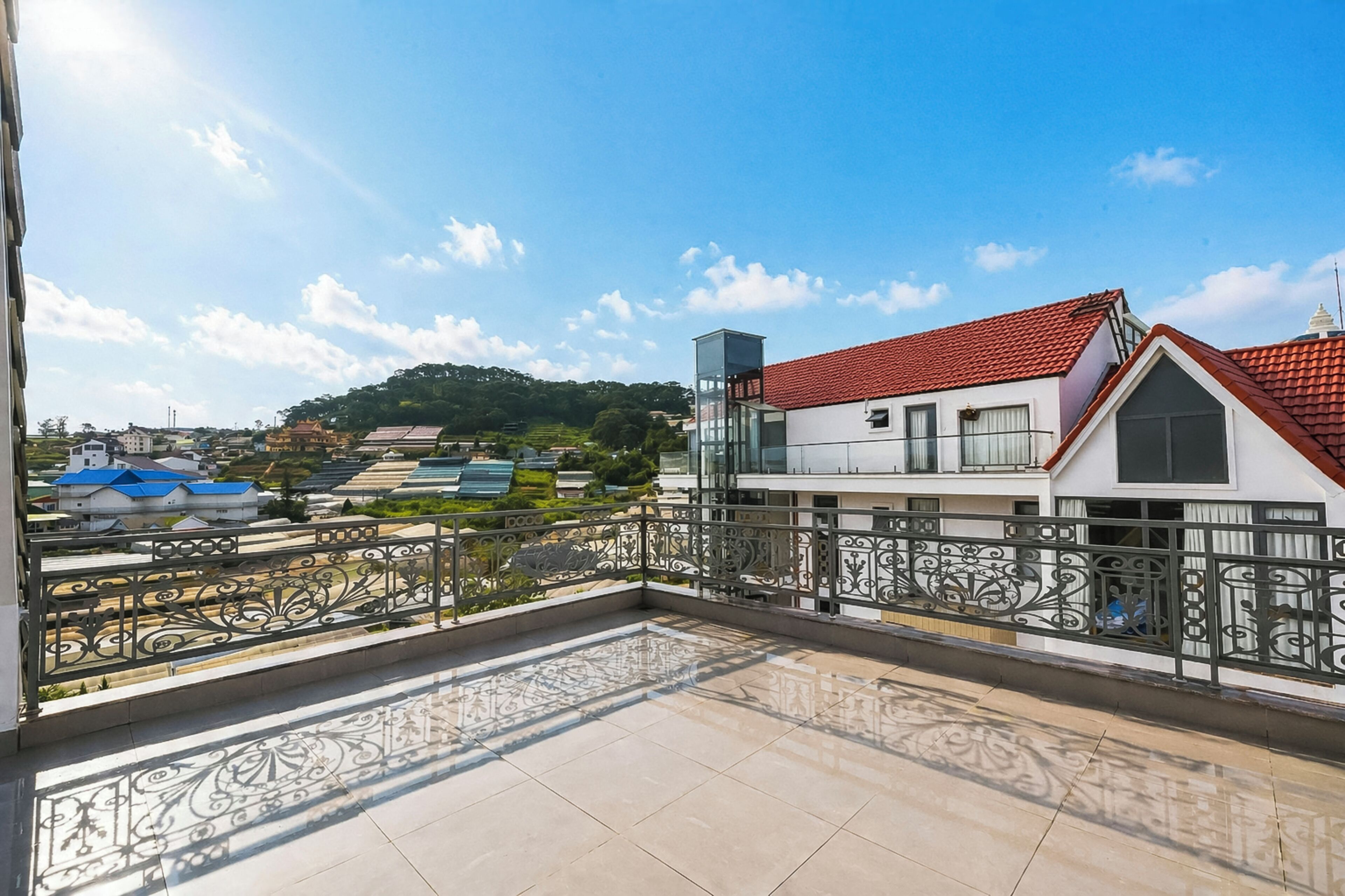 Un balcón con suelo de baldosas y barandilla decorativa, con vistas a una zona residencial con casas y una colina al fondo.