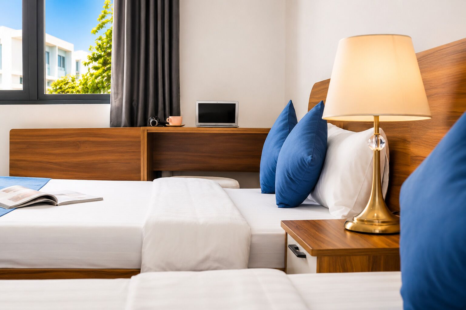 Habitación de hotel con dos camas, una mesita de noche con una lámpara, una pequeña mesa con una cámara y una taza, y una ventana con cortinas.