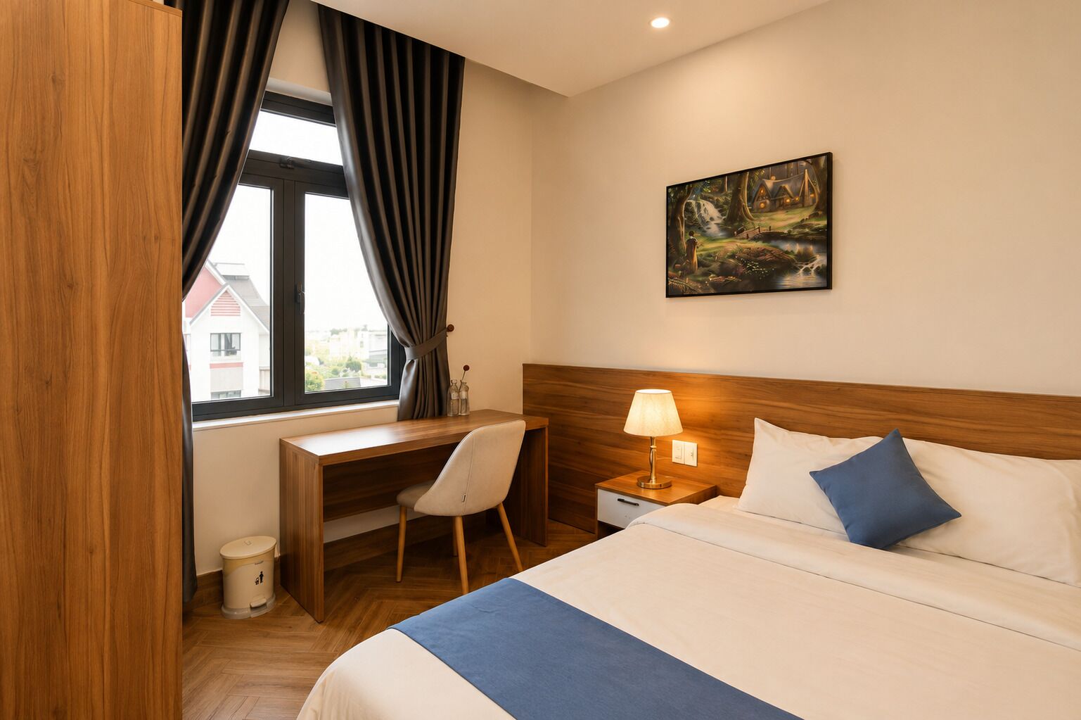 A hotel room with a bed, a desk, a chair, a nightstand with a lamp, and a painting on the wall.