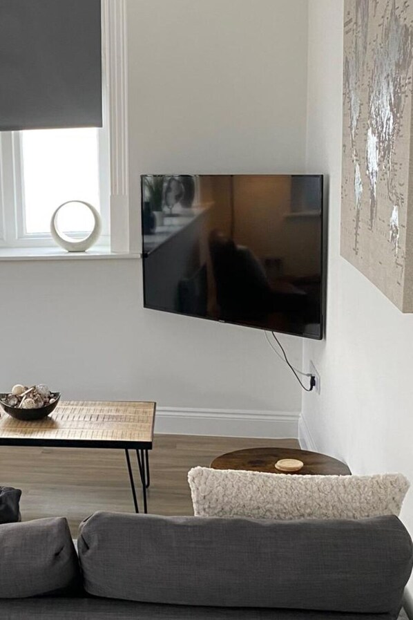 A modern living room with a flat-screen TV mounted on the wall, a round table with a bowl of eggs, and a gray sofa. - Cozy Coastal Retreat in Bournemouth (Bournemouth)