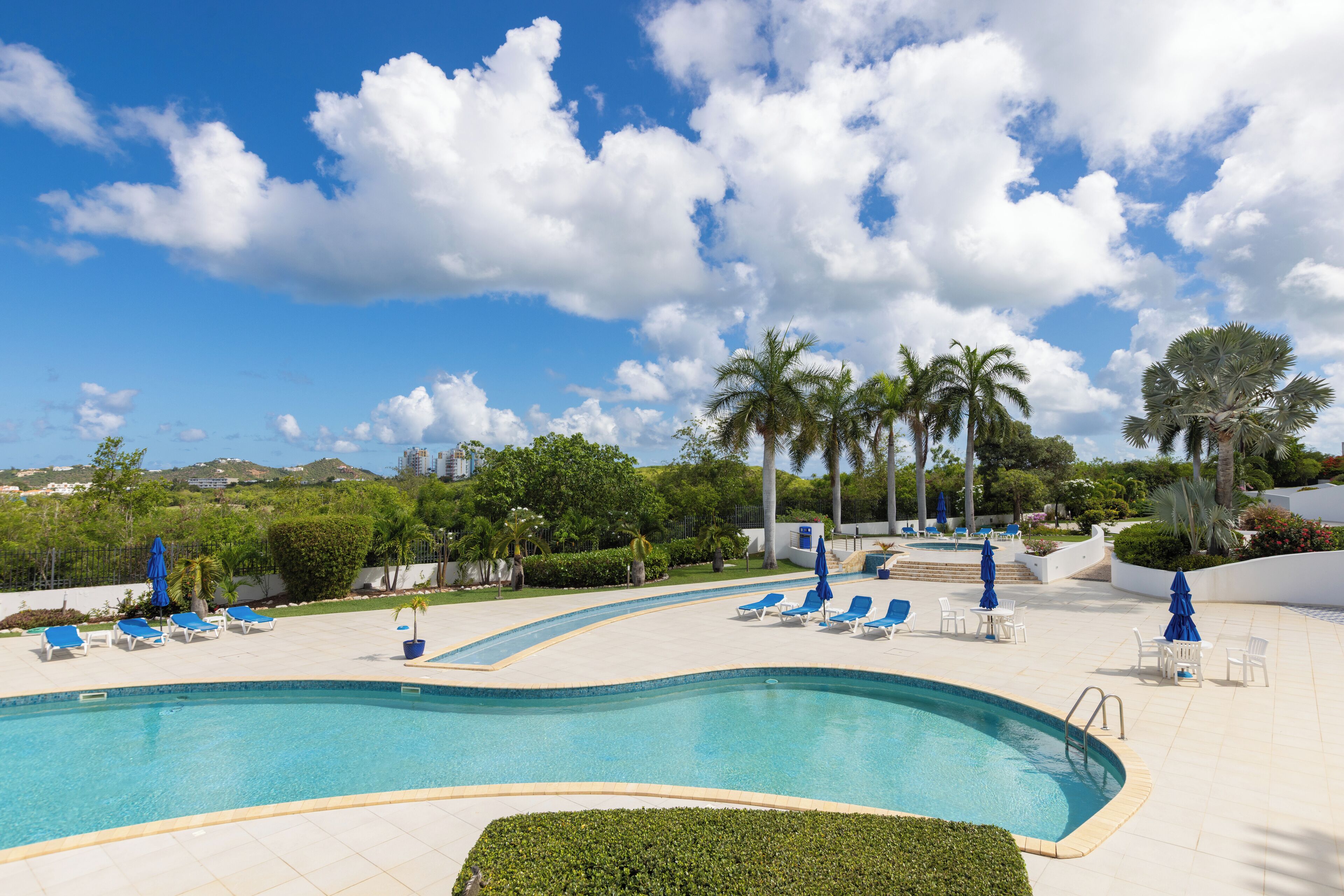 A swimming pool with lounge chairs and umbrellas, surrounded by palm trees and greenery.