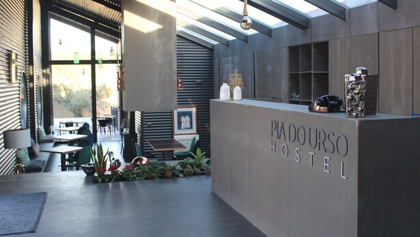 A modern hostel reception area with a concrete counter, seating area, and large windows. - Hostel Pia do Urso (Batalha)