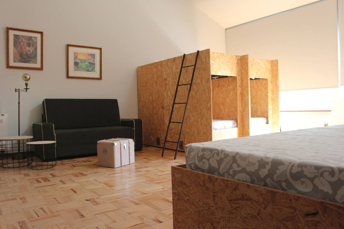 A room with a wooden floor, a sofa, a small table, a ladder, and a bed with a patterned bedspread.