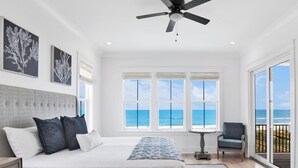 Quarto com cama, ventoinha no teto, vista para o oceano e varanda com uma cadeira.
