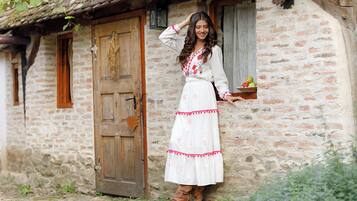 Edificio de piedra rústica con puerta y ventanas de madera, una persona vestida con traje tradicional apoyada en la pared, y un pequeño estante con fruta.