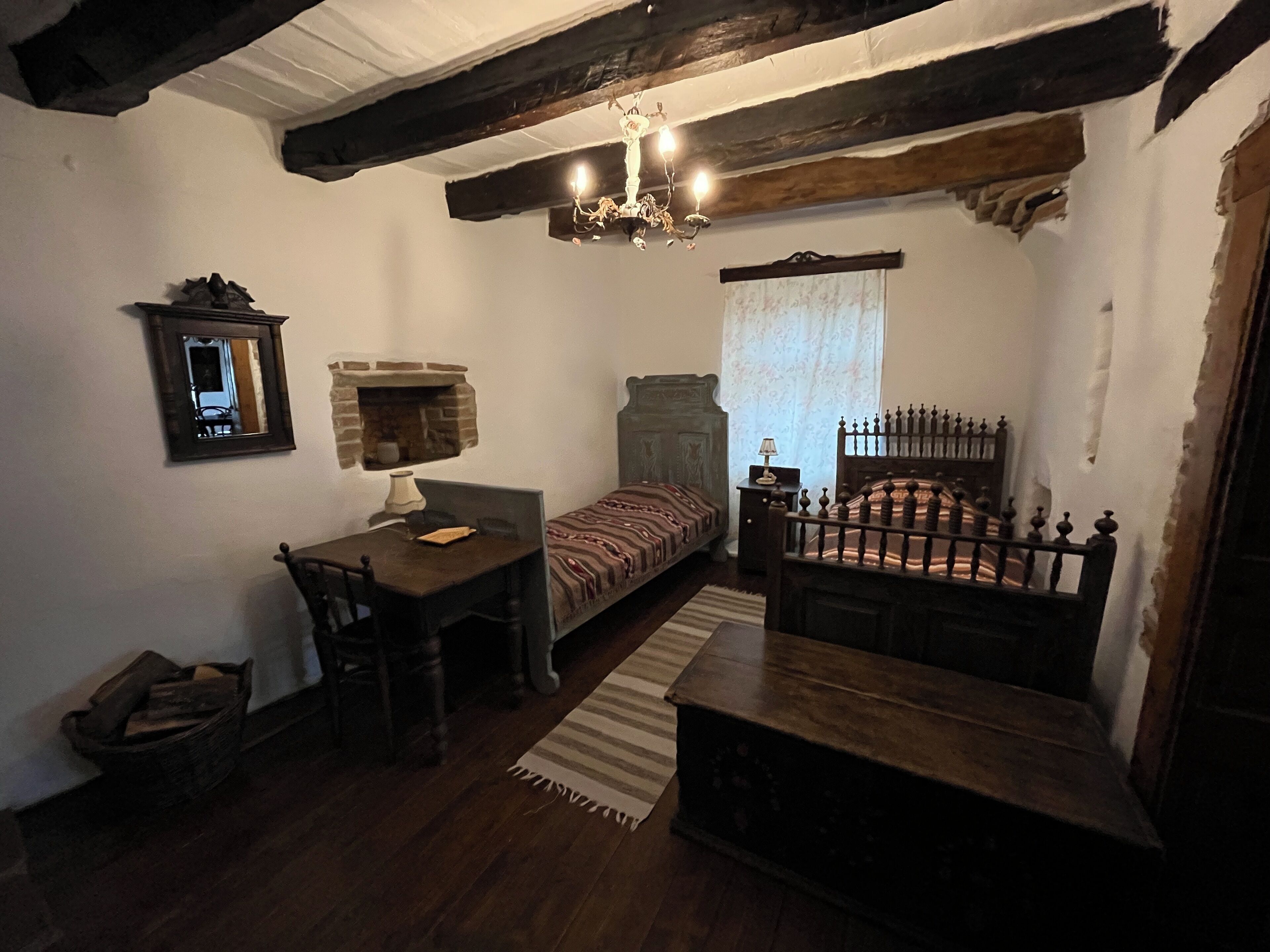 Una habitación con cama, escritorio, silla, pequeña mesa, espejo, chimenea y candelabro.