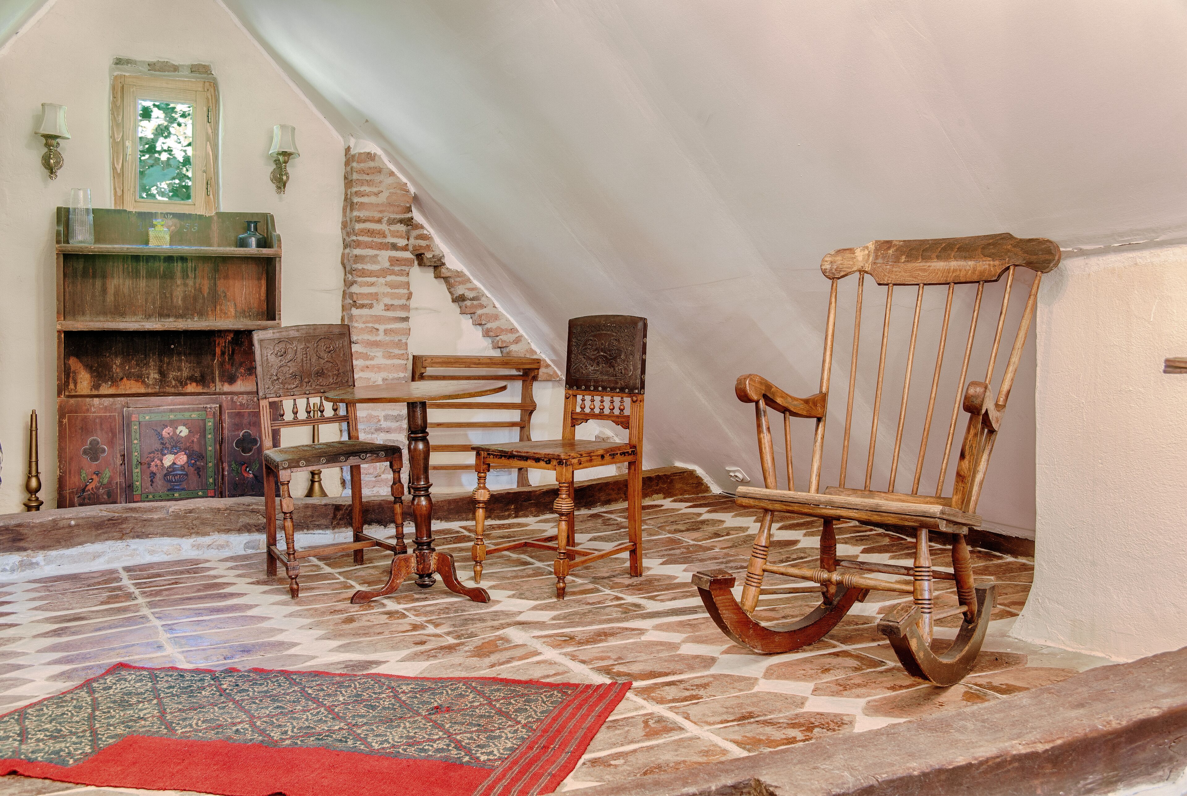 A room with a wooden rocking chair, a wooden table, a bench, and a patterned rug on a tiled floor.