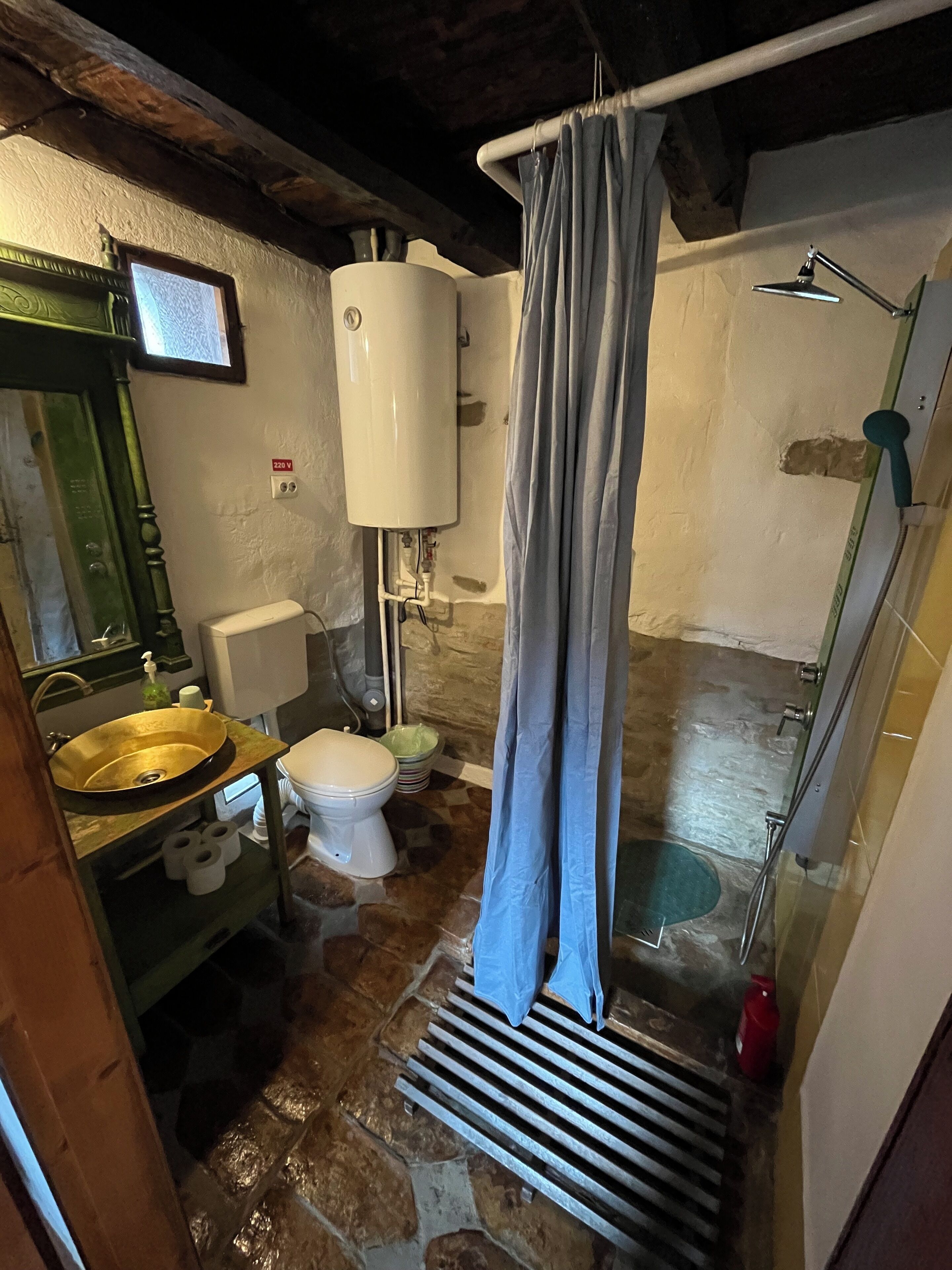 A bathroom with a stone floor, a toilet, a sink with a green countertop, a shower with a glass door, and a white water heater.