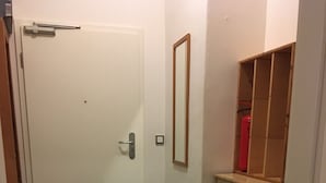 Ein schmaler Flur mit Holzboden, einer weißen Tür, einem kleinen Spiegel und einem Holzschrank mit einem roten Gegenstand darauf.