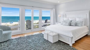 Une chambre avec une grande fenêtre donnant sur la plage, un ventilateur de plafond et un lit blanc avec des oreillers assortis.