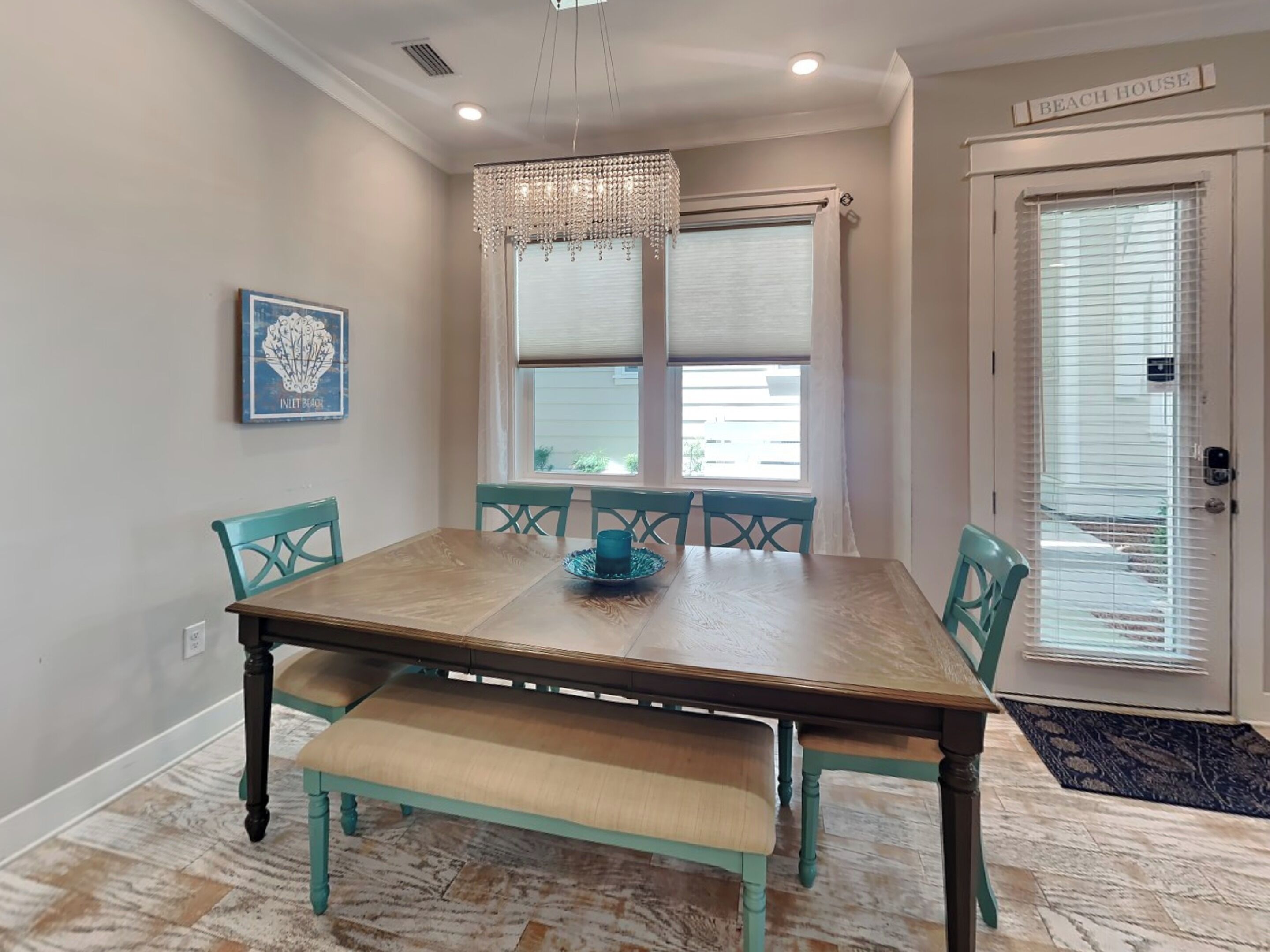 A dining room with a wooden table, teal chairs, and a bench. There is a chandelier above the table, a window with blinds, and a door labeled "Beach House".