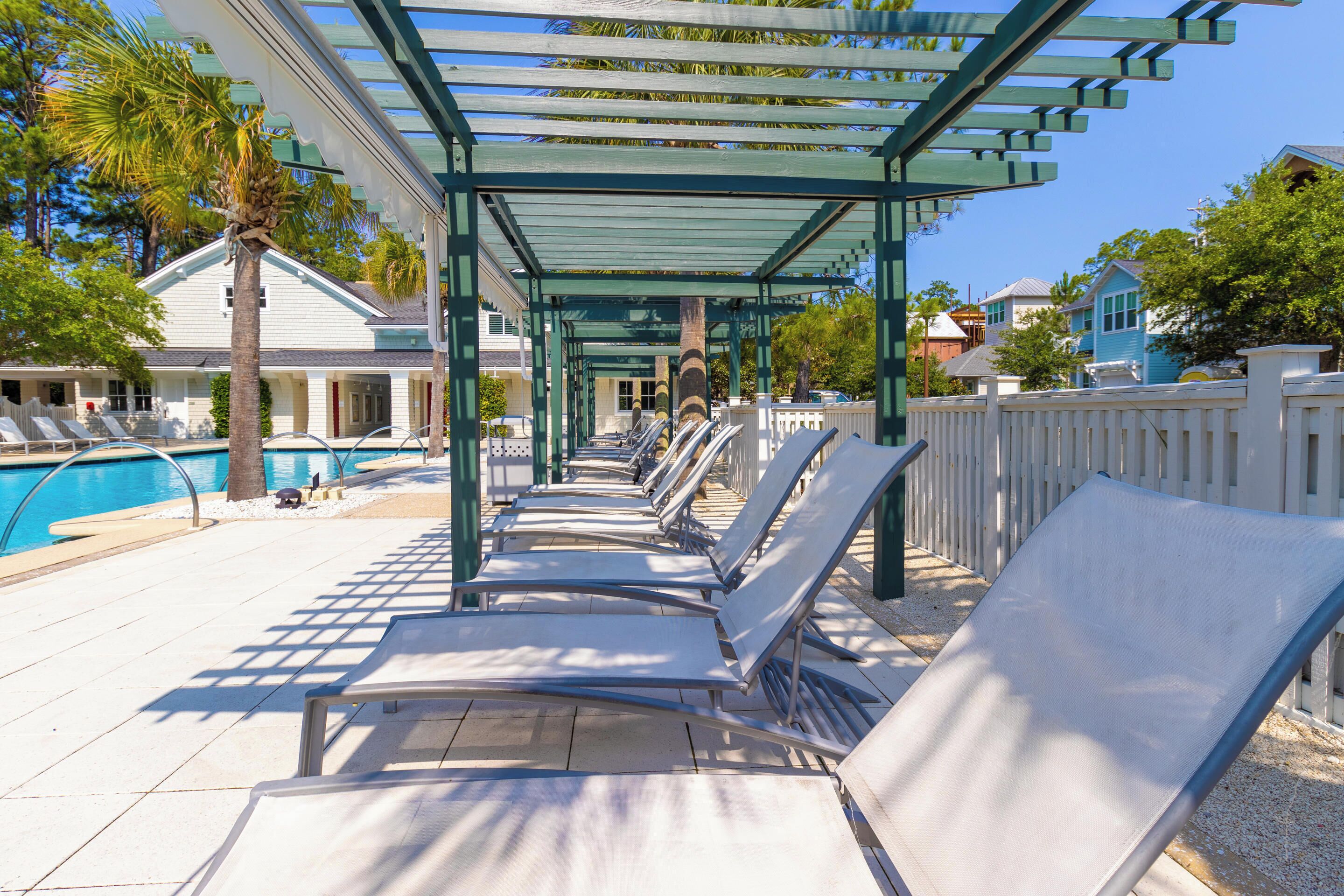 A poolside area with sun loungers under a pergola, a pool, and a white fence.
