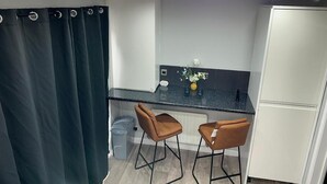 A compact kitchen with a dark countertop, two bar stools, a white refrigerator, and a small trash bin.