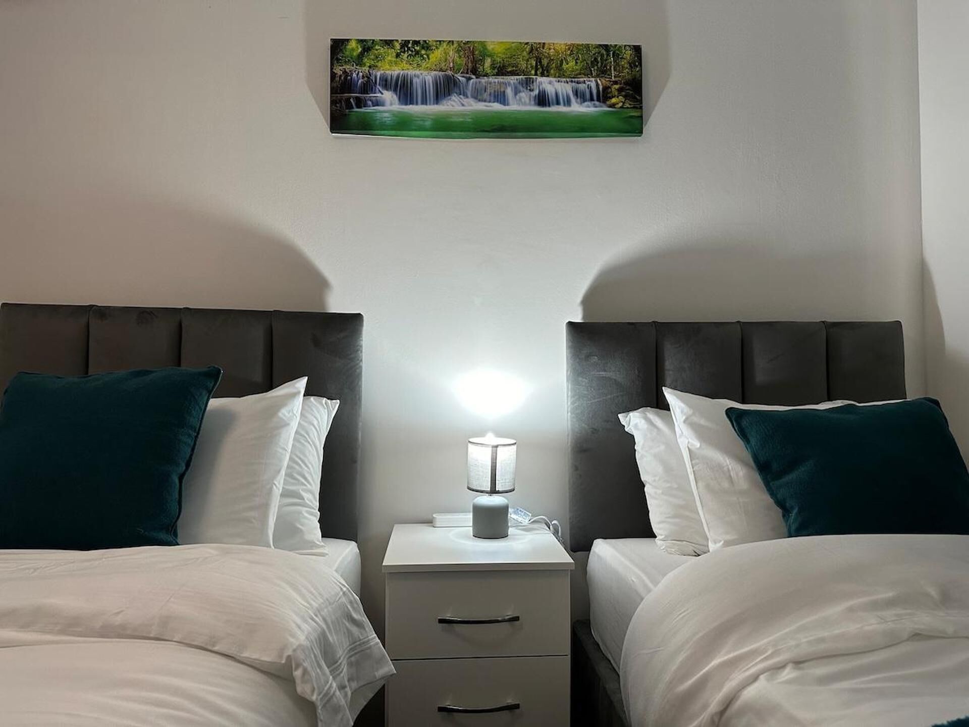 A hotel room with two beds, a nightstand with a lamp, and a painting of a waterfall on the wall.