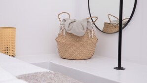 A bathroom with a woven basket on a white shelf, a round mirror with a black frame, and a textured candle holder. - Serenity Bay, Cycladic Villa with Pool and magnificent view near the beach. (Leros)