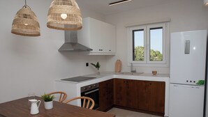 A modern kitchen with white cabinets, a wooden countertop, a built-in oven, and a white refrigerator. There are two wicker pendant lights, a wooden dining table with chairs, and a window with a view of trees. - Serenity Bay, Cycladic Villa with Pool and magnificent view near the beach. (Leros)