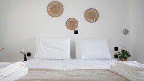 A neatly made bed with white linens, a patterned throw blanket, and a stack of folded towels. There are three woven wall hangings above the bed, a small potted plant, and a wall-mounted lamp. - Serenity Bay, Cycladic Villa with Pool and magnificent view near the beach. (Leros)