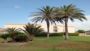 A building with a flat roof and a beige facade, surrounded by a well-maintained lawn and palm trees. - Faro's Retamar Cuddly Nest for 2 (Almería)