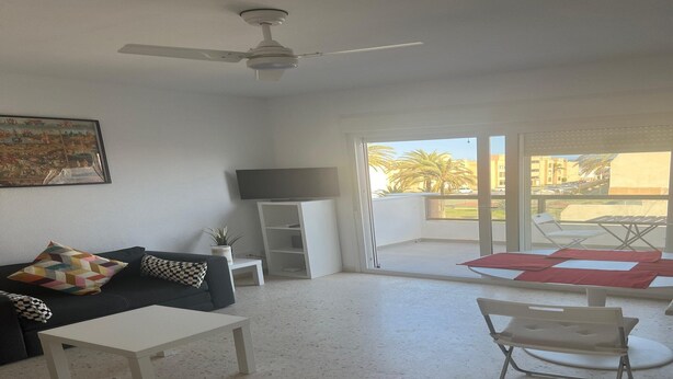 A living room with a black sofa, white coffee table, and a TV stand. There is a ceiling fan, a window with a view of palm trees, and a balcony with a red and white mat. - Faro's Retamar Cuddly Nest for 2 (Almería)