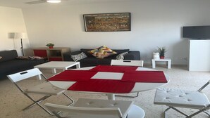 A living room with a round table, white chairs, a black sofa, a ceiling fan, a painting on the wall, and a television on a stand. - Faro's Retamar Cuddly Nest for 2 (Almería)