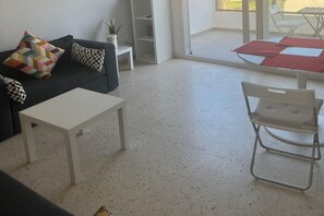 A living room with a sofa, a coffee table, a metal chair, and a glass door leading to an outdoor area. - Faro's Retamar Cuddly Nest for 2 (Almería)