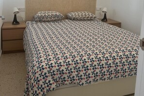 A bed with a patterned bedspread, two pillows, a bedside lamp, and a wooden nightstand. - Faro's Retamar Cuddly Nest for 2 (Almería)