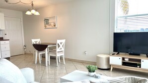Een moderne woonkamer met een bank, een eettafel met stoelen, een televisie op een dressoir en een raam met jaloezieën.
