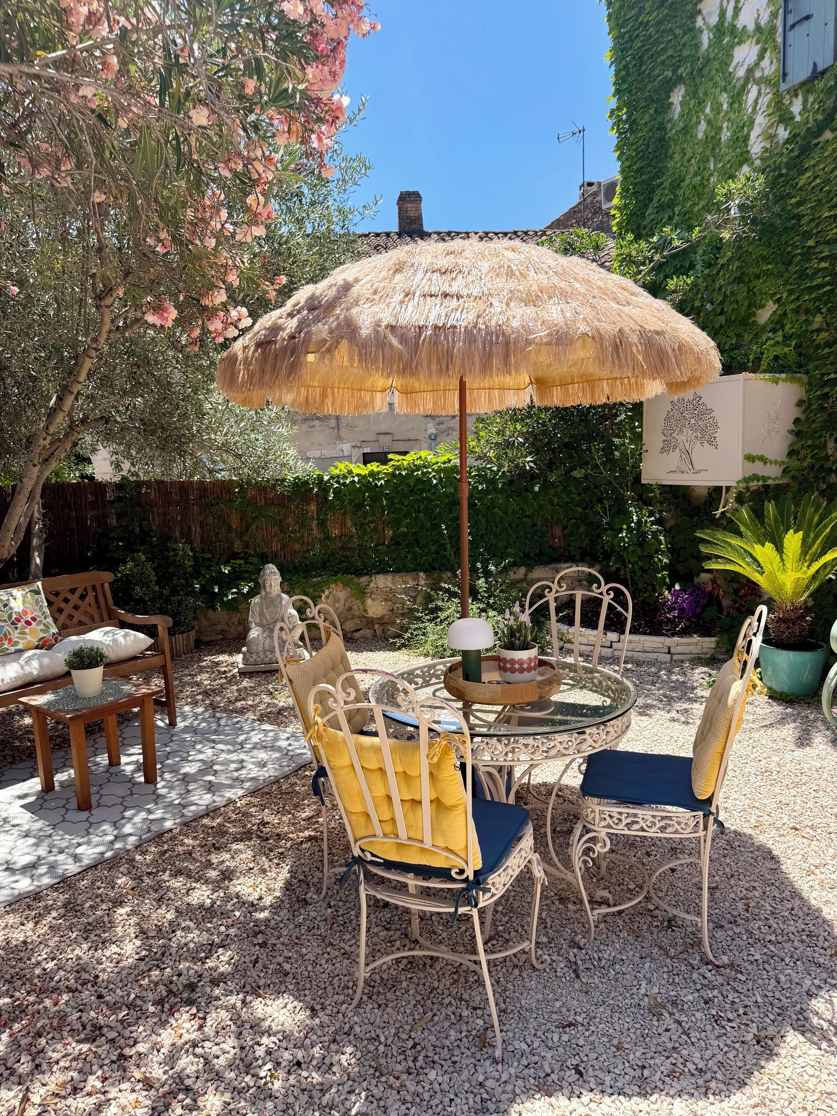 An outdoor seating area with a thatched umbrella, wicker chairs, a table, and a Buddha statue.