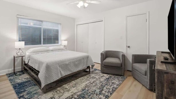 Ein Schlafzimmer mit einem Bett, zwei Stühlen, einem Fernseher, einem Deckenventilator und einem Fenster mit Jalousien.